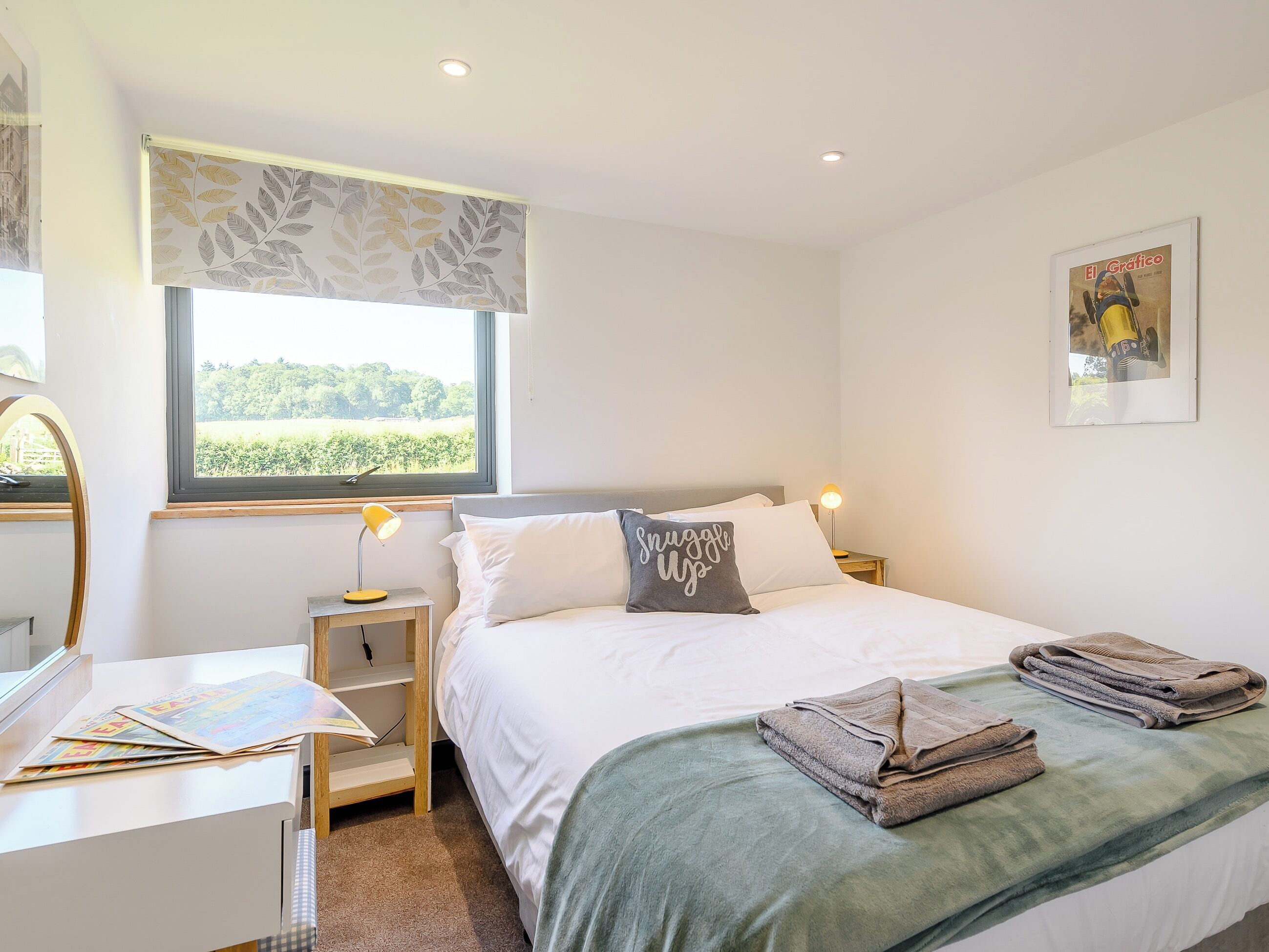 A bedroom with a bed, a desk, a lamp, a window with a view of greenery, and a framed picture on the wall.