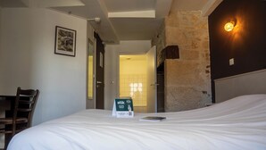 A hotel room with a bed, a chair, a framed picture on the wall, a sign for a 5.5% discount, and a stone wall. - Logis Hôtel EBEN (Egletons)