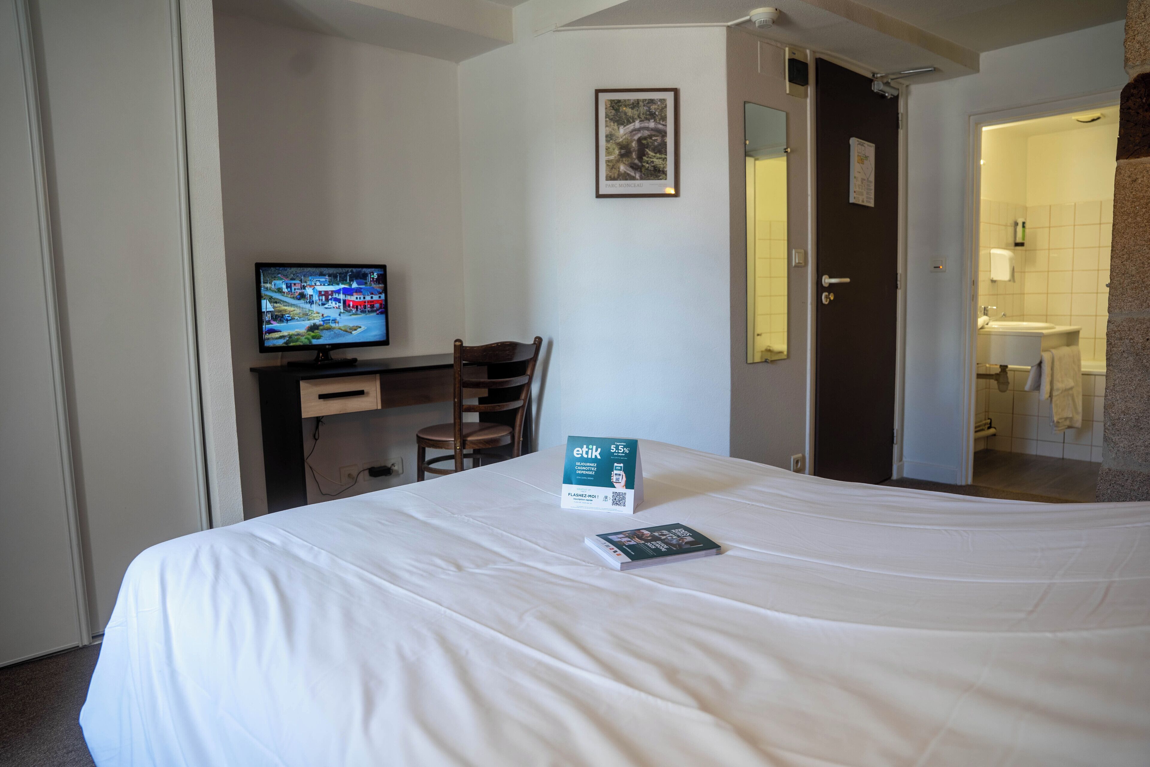 Una camera d'albergo con un letto, una scrivania con un computer, una televisione e un bagno visibile attraverso una porta aperta.