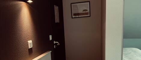 Habitación de hotel con cama, aire acondicionado de pared, un cuadro enmarcado y una ventana con cortina blanca.