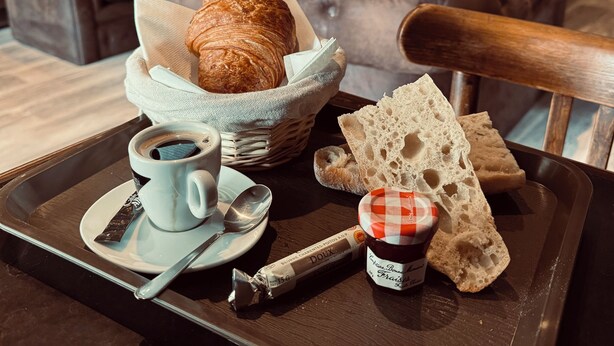 A breakfast tray with croissants, bread, jam, coffee, and a pastry on a tray in a cozy room with leather sofas and a plant. - Logis Hôtel EBEN (Egletons)