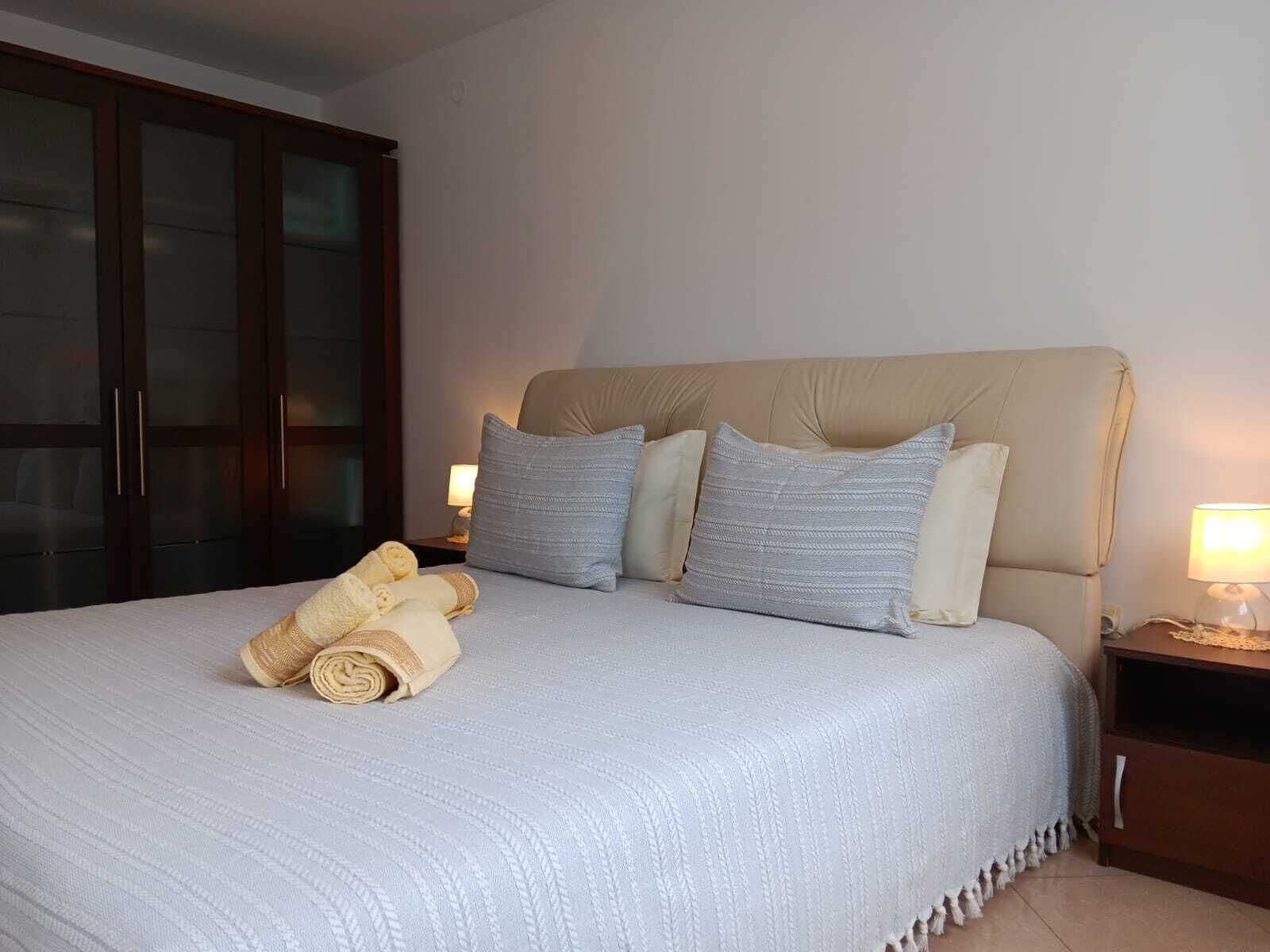 A neatly made bed with a beige headboard, white bedding, and rolled towels on the side.