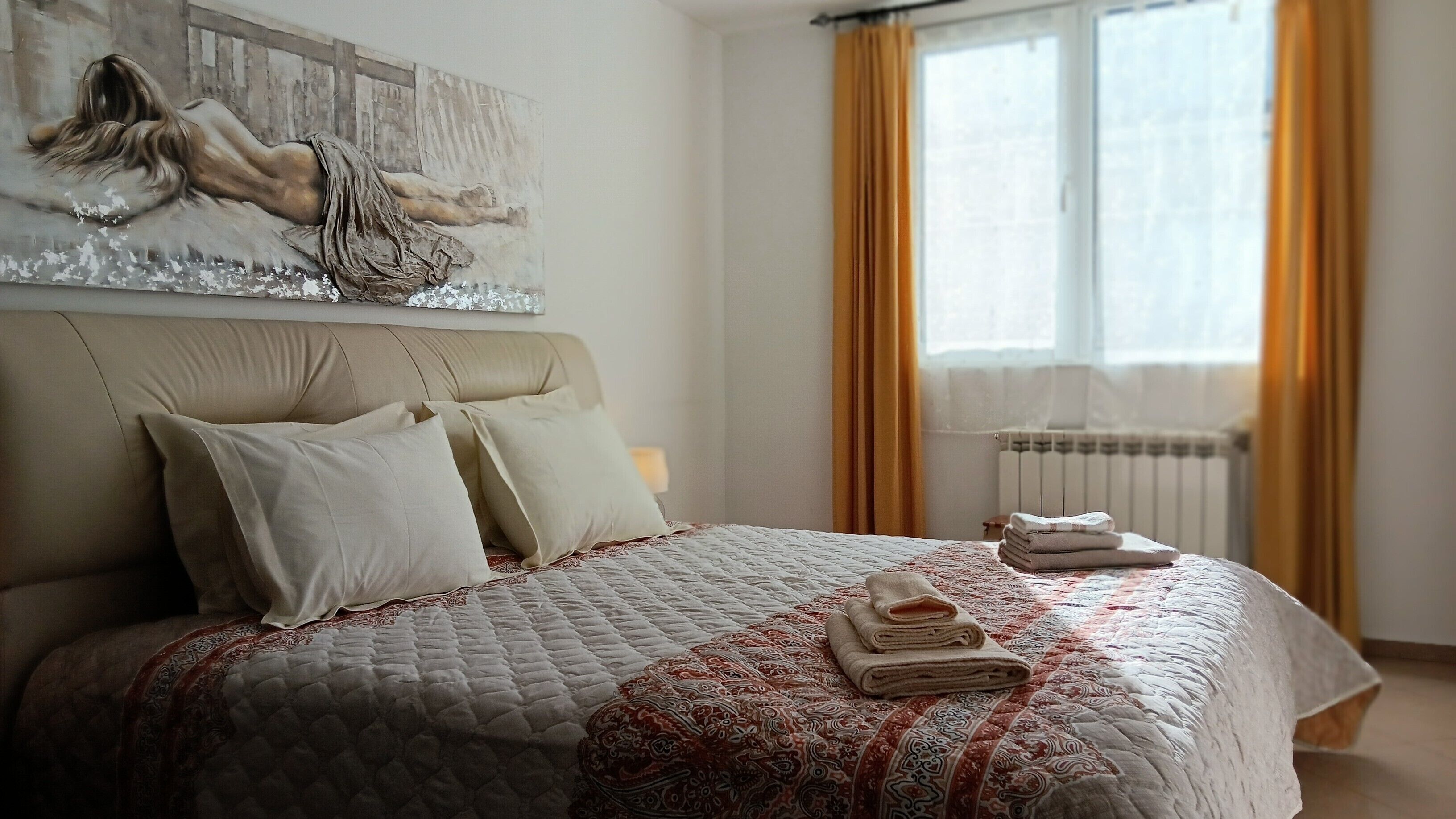 Une chambre à coucher avec un grand lit, une tête de lit, un tableau, une fenêtre avec des rideaux et un radiateur.