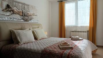 Une chambre à coucher avec un grand lit, une tête de lit, un tableau, une fenêtre avec des rideaux et un radiateur.