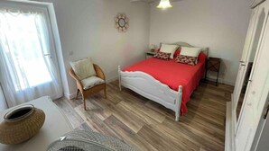 Un dormitorio con una cama blanca, ropa de cama roja, una silla de mimbre, una mesita y una lámpara colgante.