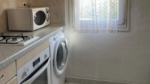 Cocina compacta con microondas, horno y lavadora. Se aprecia una ventana con cortina estampada y una ventana pequeña con marco blanco.