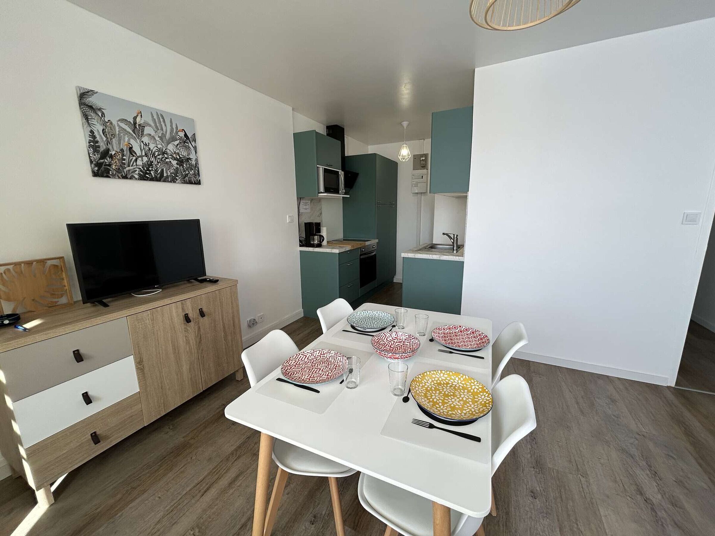 Una cocina moderna con armarios azul verdoso, un microondas y un fregadero. Hay una mesa de comedor con sillas blancas y platos. Un televisor de pantalla plana está sobre un mueble de madera.