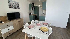 A modern kitchen with teal cabinets, a microwave, and a sink. There is a dining table with white chairs and plates. A flat-screen TV is on a wooden cabinet. - Beachside 2-Room Apartment with Private Garage in Saint-Hilaire-de-Riez (Saint-Hilaire-de-Riez)