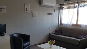 Ein Wohnzimmer mit einer Couch, einem schwarzen Sessel, einem weißen Couchtisch mit Blumenvase und einem Fernseher auf einem Schrank.