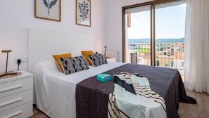 A bedroom with a large bed, two pillows, a bedside table, a lamp, and a view of the city through a large window. - Modern Seaside Apartment, Sant Antoni (San Antonio de Calonge)