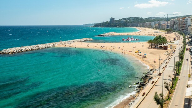 A coastal beach with clear turquoise waters, a rocky breakwater, and a promenade lined with buildings and palm trees. - Modern Seaside Apartment, Sant Antoni (San Antonio de Calonge)