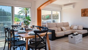 A modern living room with a dining area, large windows, and a balcony with a view of greenery. - Family-Friendly House with Pool in Calonge, 5min to Beach (Calonge)