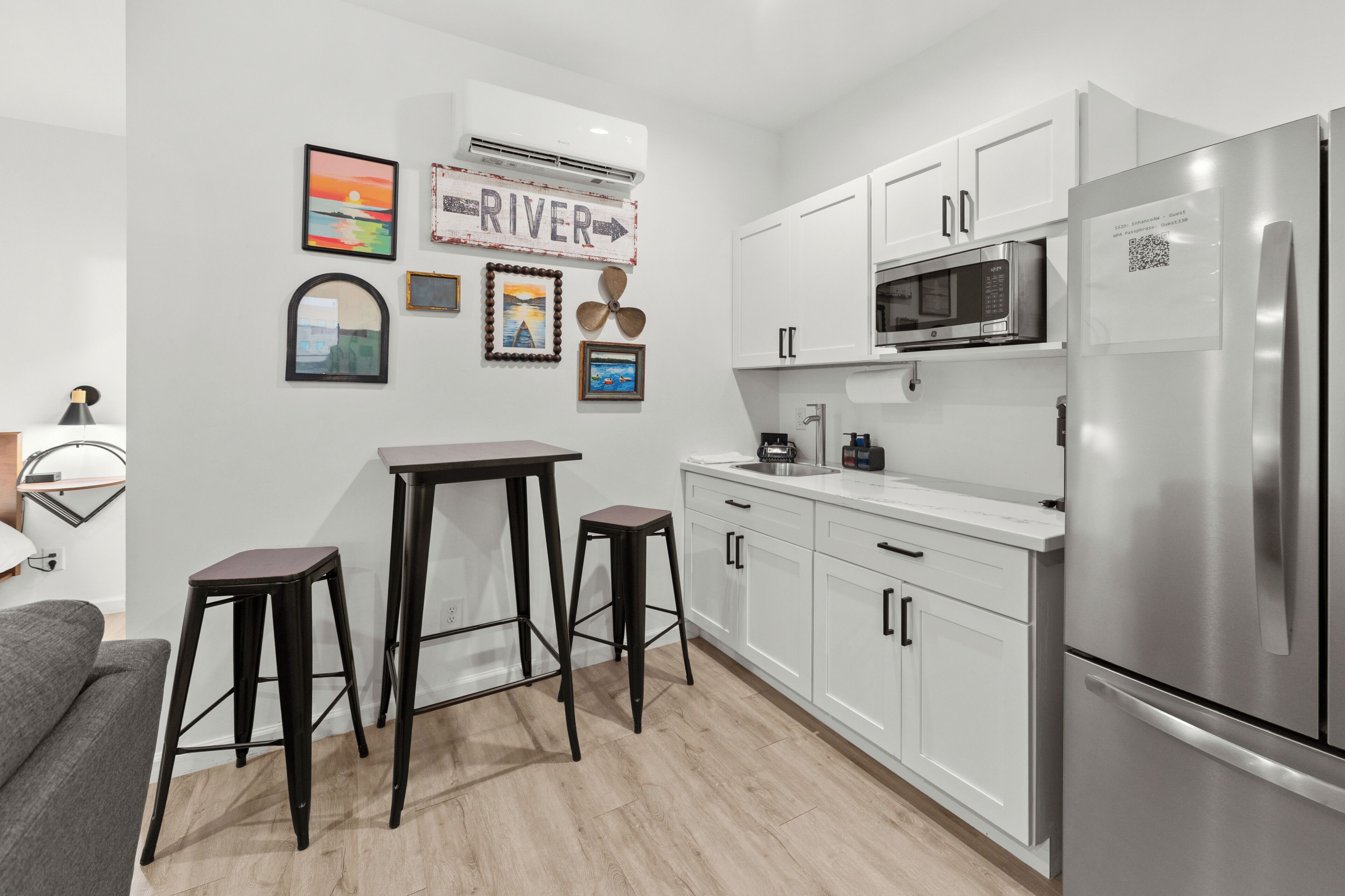 Eine moderne Küche mit weißen Schränken, einem Edelstahl-Kühlschrank, einer Mikrowelle und einem Spülbecken. Zwei Hocker und ein Tisch stehen bereit. Auf der Wand ist ein Kunstwerk angebracht.