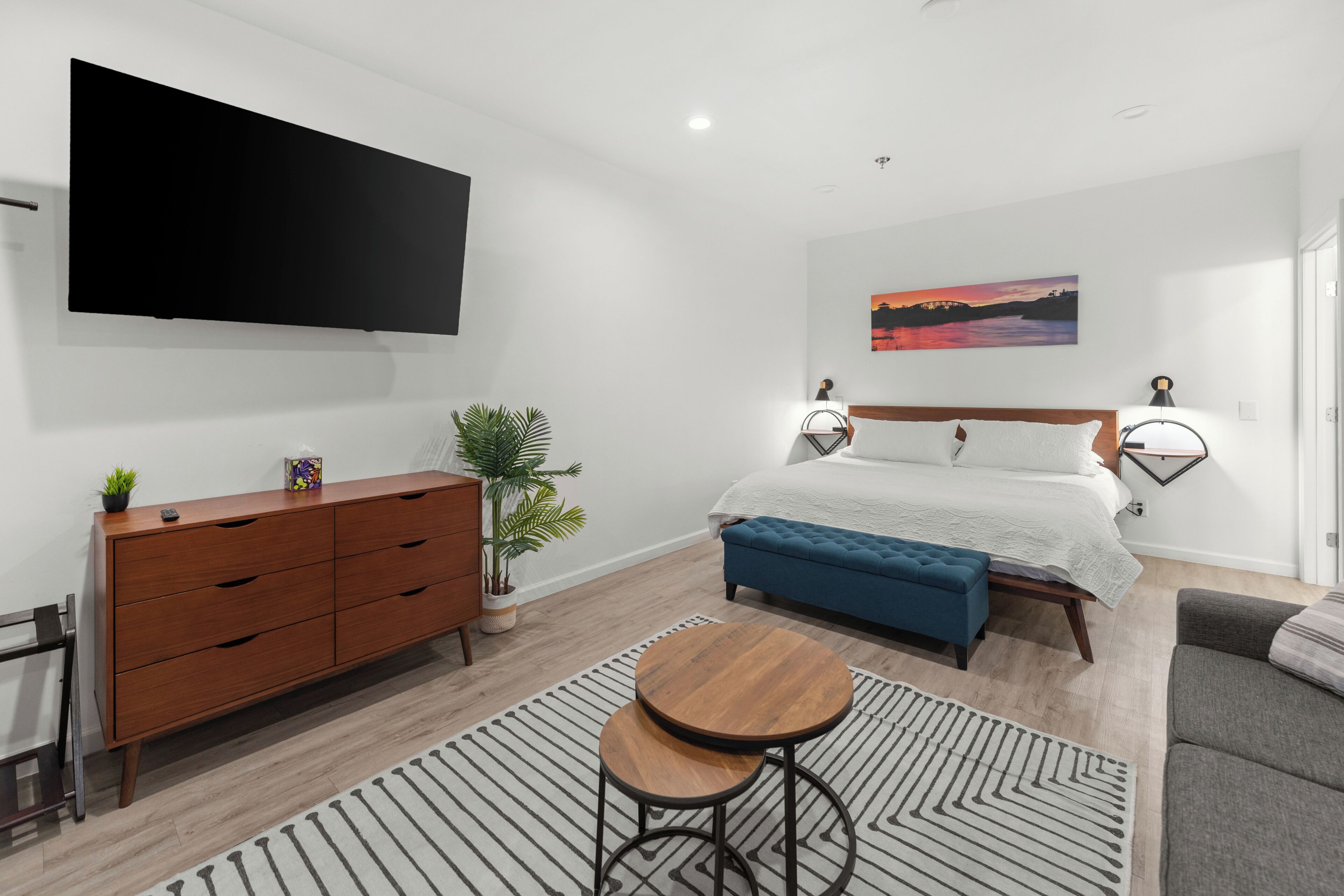 Ein modernes Schlafzimmer mit Bett, Nachttischlampen, Fernseher, Kommode, Pflanze und Sofa.