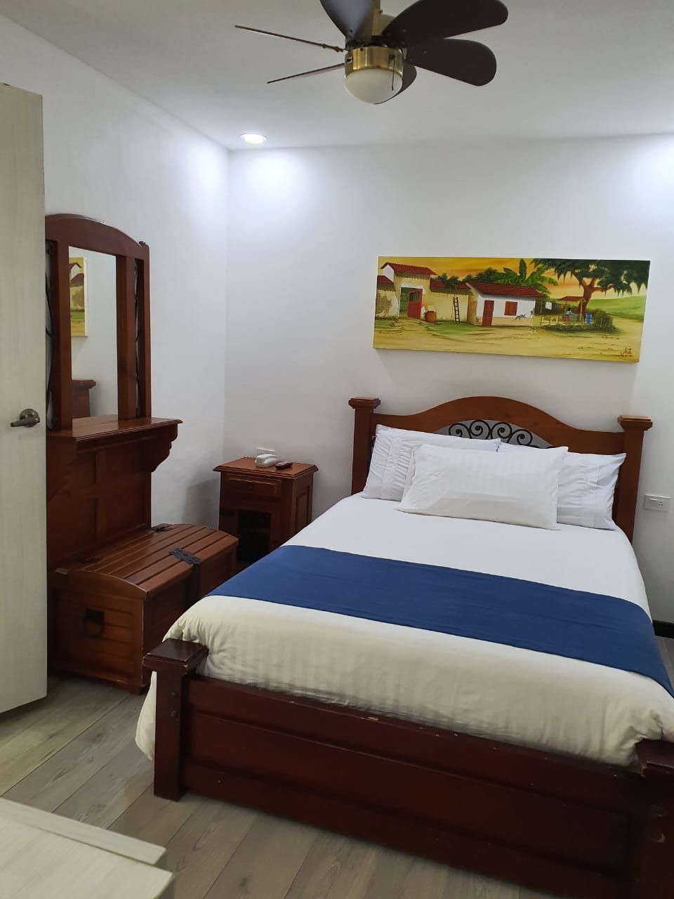 Un dormitorio con una cama, una mesita de noche, un banco de madera, un ventilador de techo y un cuadro en la pared.