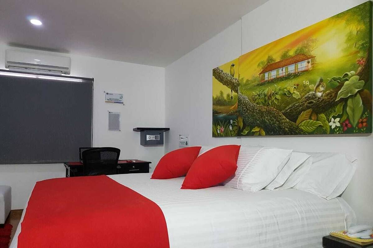 Habitación de hotel con cama, almohadas rojas y blancas, escritorio con silla, una mesita y un cuadro en la pared.