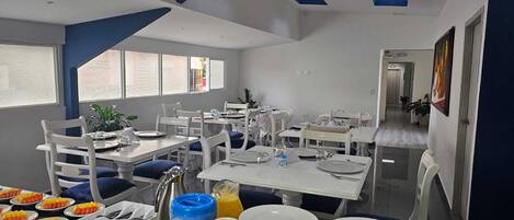 A modern dining area with white furniture, blue accents, and a view of the street through large windows.