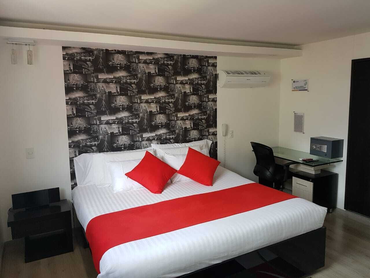 A hotel room with a large bed, red pillows, a desk with a computer, and a cityscape wallpaper.