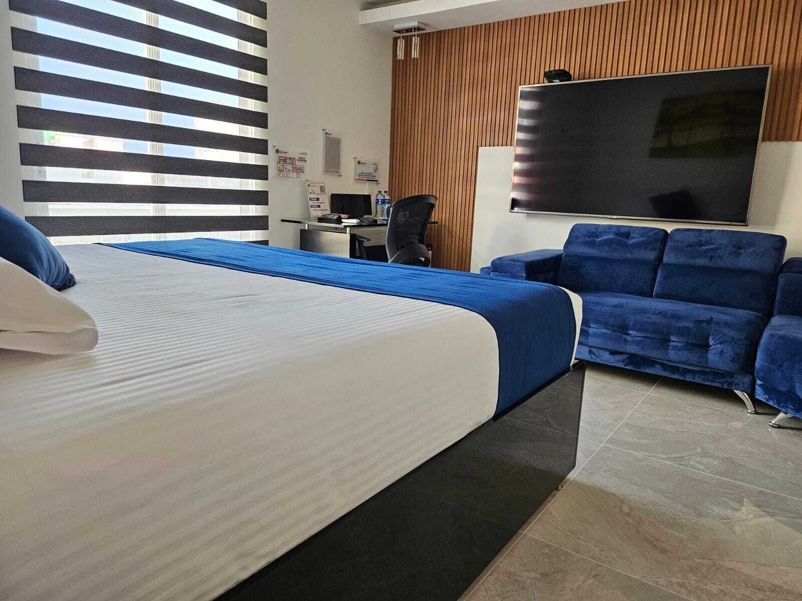 A hotel room with a bed, a blue sofa, a desk with a computer, and a TV on the wall.