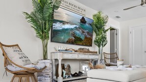 A living room with a large TV mounted on the wall, a white sofa, a wicker chair, and a white console table with decorative items. - Dolphin Bay Bungalow – Waterfront Pool, Kayaks, & Walk to Indian Rocks Beach (Indian Rocks Beach)