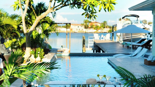 A pool area with lounge chairs, a small table with a hat and drink, and a view of the water and boats in the background. - Dolphin Bay Bungalow – Waterfront Pool, Kayaks, & Walk to Indian Rocks Beach (Indian Rocks Beach)