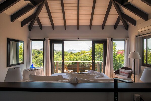 A bedroom with a bed, a nightstand, a lamp, and a view of greenery and mountains through large windows.