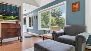 Une pièce avec une grande fenêtre, une commode en bois, un fauteuil gris et un tableau accroché au mur.
