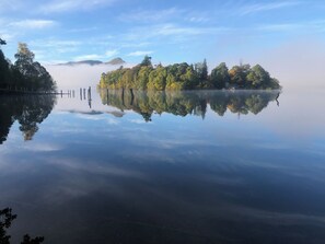 A calm lake with a clear reflection of trees and a misty island in the distance.