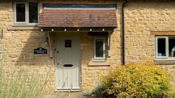 A stone cottage with a white door, a small porch, and a sign reading "HoneySuckle Cottage."
