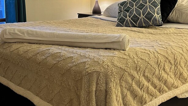 A neatly made bed with a fluffy white blanket, a patterned pillow, and a bedside lamp on a nightstand. - Fast Track SCL AirPort (Santiago)