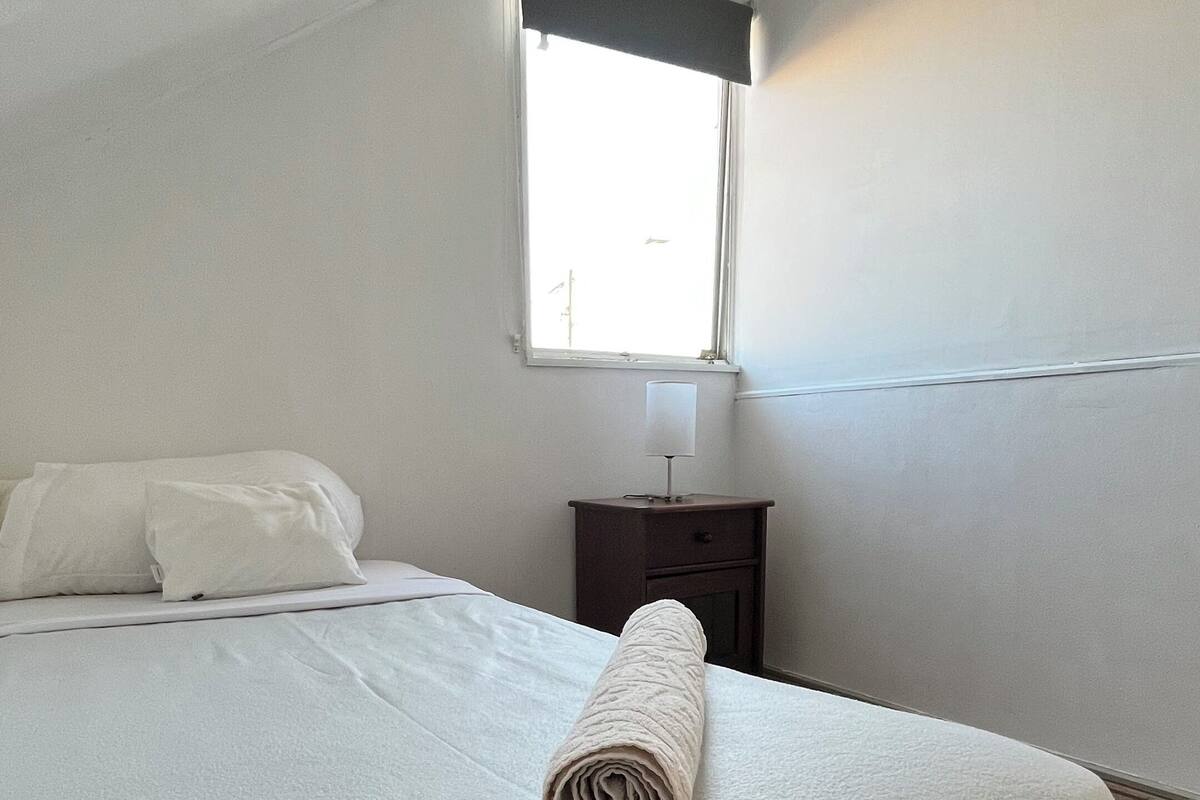 A neatly made bed with a rolled towel, a bedside table with a lamp, and a window with a view of the outside.