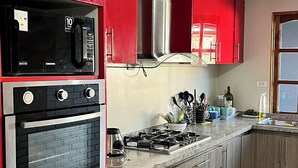 A kitchen with red cabinets, a stainless steel range, and a microwave. There are wooden cabinets below, a gas stove, and a tiled floor with a black border. - Fast Track SCL AirPort (Santiago)