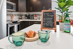A kitchen counter with a chalkboard menu, two green glass cups, a croissant, and a bottle of Pellegrino water. - Tropical Toucan Retreat w/ Pool (Pompano Beach)