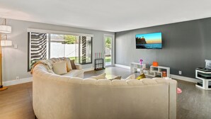 A modern living room with a large sofa, a flat-screen TV mounted on the wall, and a view of the outdoors through large windows. - Tropical Toucan Retreat w/ Pool (Pompano Beach)