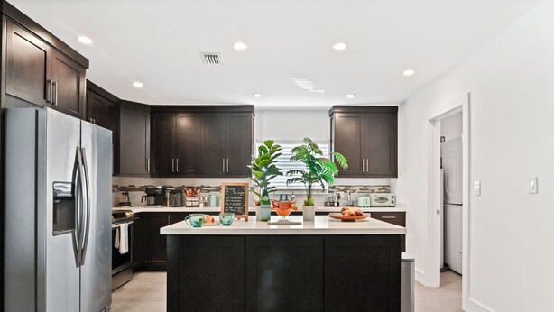 A modern kitchen with dark cabinetry, a stainless steel refrigerator, a central island with a plant, and a chalkboard menu. - Tropical Toucan Retreat w/ Pool (Pompano Beach)