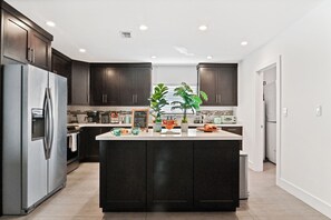 A modern kitchen with dark cabinetry, a stainless steel refrigerator, a central island with a plant, and a chalkboard menu. - Tropical Toucan Retreat w/ Pool (Pompano Beach)