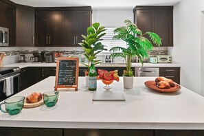 A modern kitchen with dark wood cabinets, a white countertop, and various appliances. There is a chalkboard sign, a green plant, and a plate of pastries on the counter. - Tropical Toucan Retreat w/ Pool (Pompano Beach)