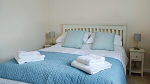 A neatly made bed with a quilted blue bedspread and white pillows, two bedside lamps, and a white nightstand with a drawer.