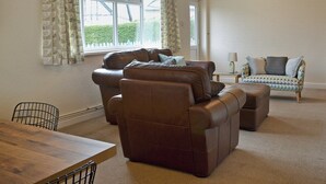 A living room with a brown leather sofa, a patterned armchair, a wooden coffee table, and a dining table with chairs.