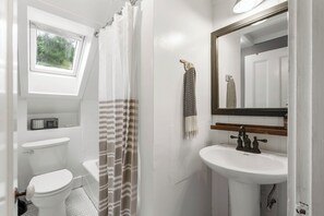 A bathroom with a white toilet, a shower with a striped curtain, a sink with a mirror, and a window above the toilet.