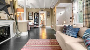 A spacious living room with a fireplace, a sofa with patterned cushions, a bookshelf, and a large window with curtains. - 2BR Historic Marblehead Home+Minutes to Beach (Marblehead)