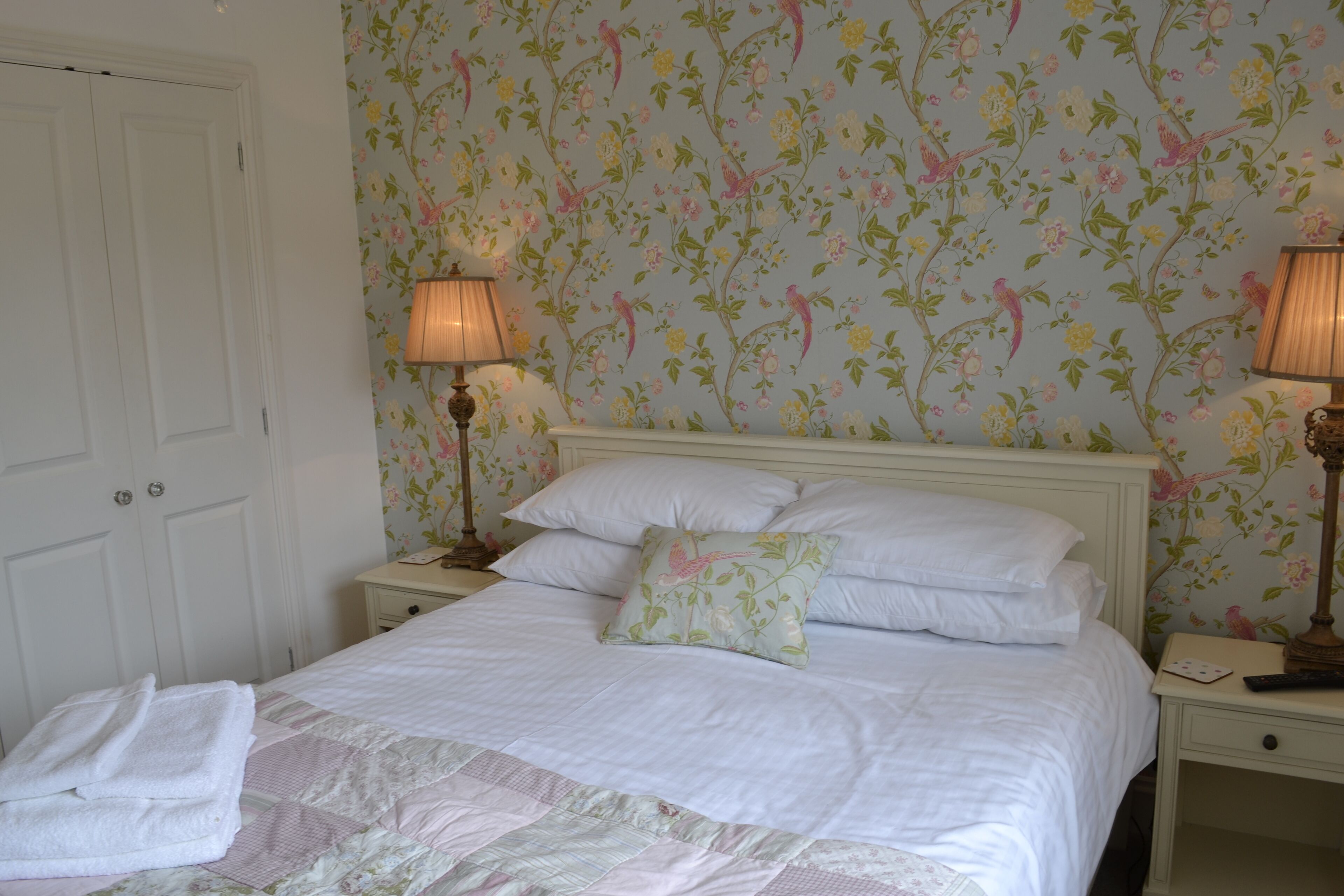 Un lit bien fait, avec du linge de lit blanc et une couverture à motifs floraux, flanqué de deux lampes de chevet et d ’une table de nuit avec une lampe.