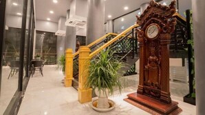 Un vestíbulo con piso de mármol, una escalera con barandilla amarilla, un gran reloj de madera y plantas en macetas.