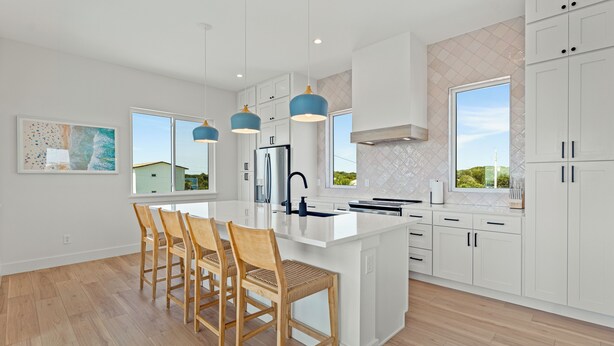 A modern kitchen with white cabinetry, a central island with bar stools, pendant lighting, and large windows offering views of the outdoors.