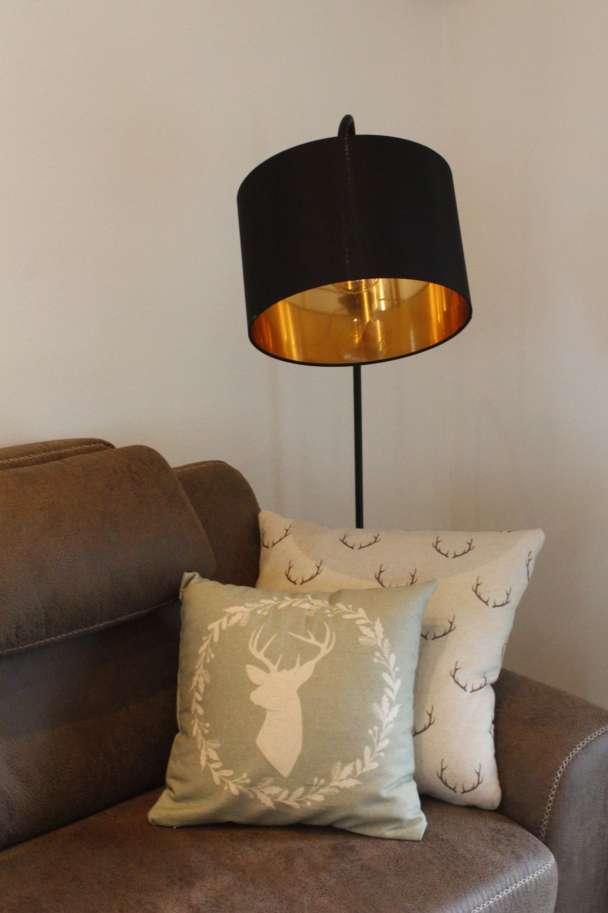 A brown leather sofa with two decorative pillows, one with a deer motif and the other with antlers. A black floor lamp with a gold shade is positioned behind the sofa.