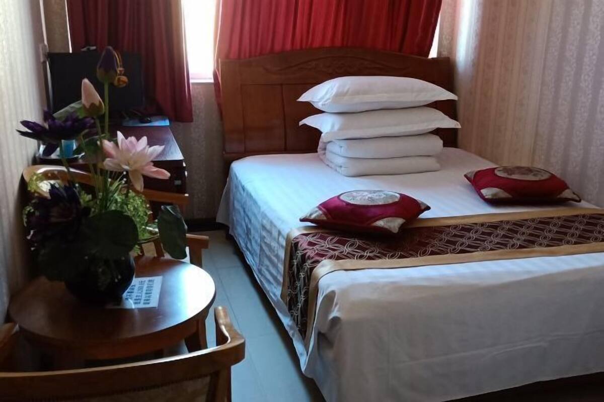 A hotel room with a bed, pillows, a bedside table with a vase of flowers, and a desk with a computer.