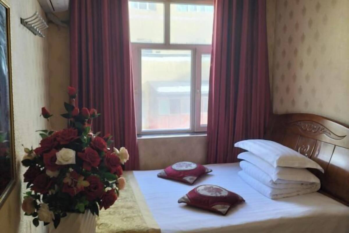 A hotel room with a bed, a window with curtains, and a vase of flowers.