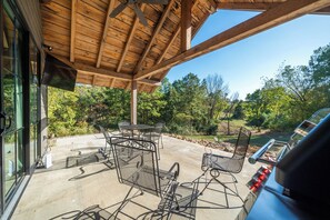 A covered patio area with outdoor furniture, a grill, and a view of a wooded landscape.