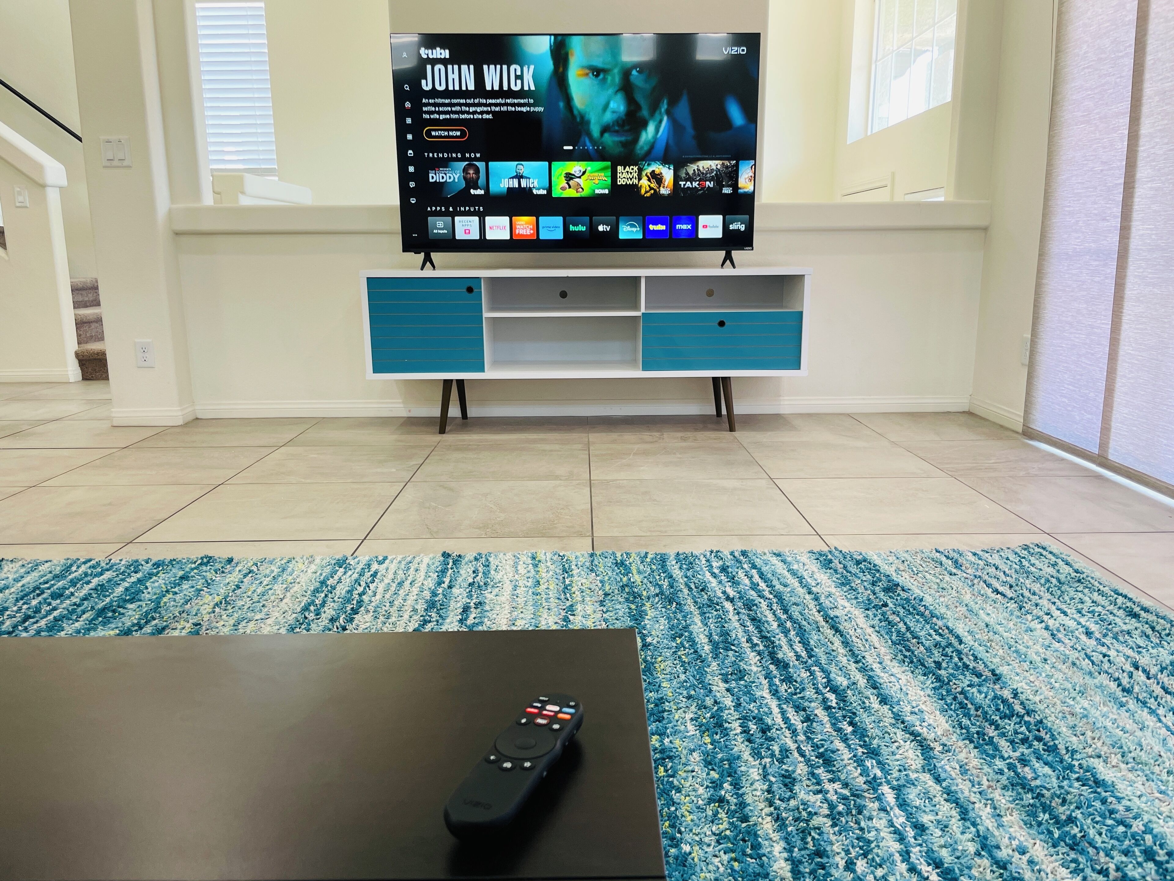 A living room with a TV mounted on the wall, a blue and white rug, and a black coffee table with a remote control.