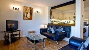 Una sala de estar moderna con un televisor de pantalla plana, una mesa de centro de vidrio, un sofá con almohadones decorativos y una cocina visible a través de un vano abierto.
